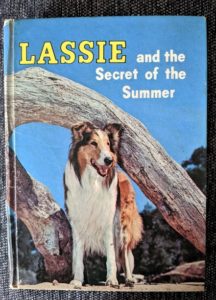 front cover of a book saying "Lassie and the Secret of the Summer," with a picture of Lassie (a sable and white Rough Collie with a white facial blaze) standing in front of a driftwood tree trunk