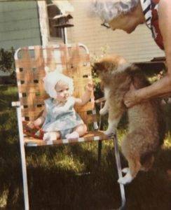 Baby wearing a bonnet and dress sitting in a chair, reaching to pet a puppy being held up to her.