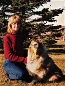 Veleda and blue merle Rough Collie Trinket seated outside before a pine tree.