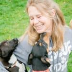 A blond-haired, smiling woman outside being licked in the face by one dog and nuzzled by another.