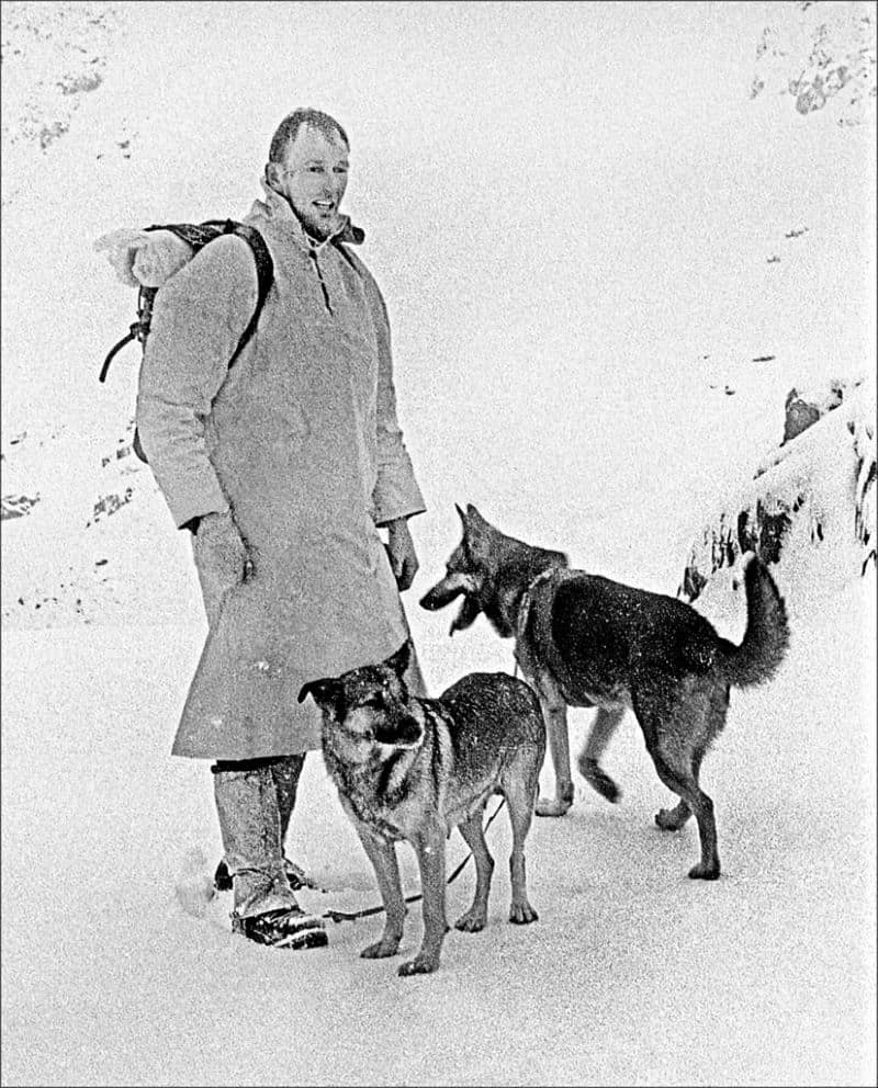 Black and white photo of a tall man in a winter coat standing with 2 search dogs that appear to be German Shepherds