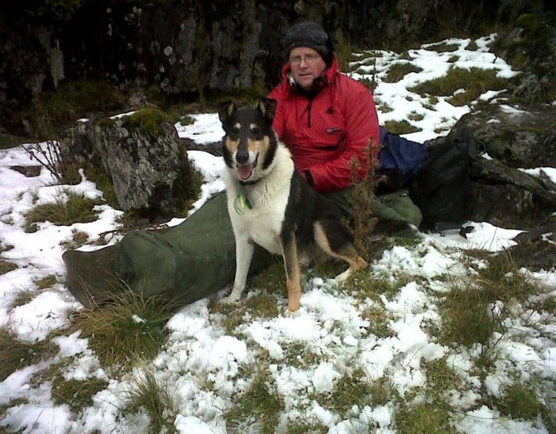 Finn sits in the snow beside a man sitting up in a sleeping bag