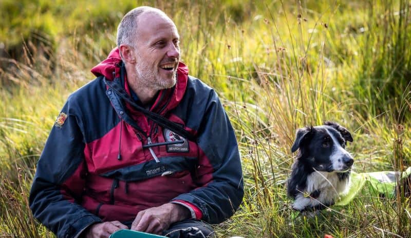 a smiling handler sits on the ground next to his border collie search dog