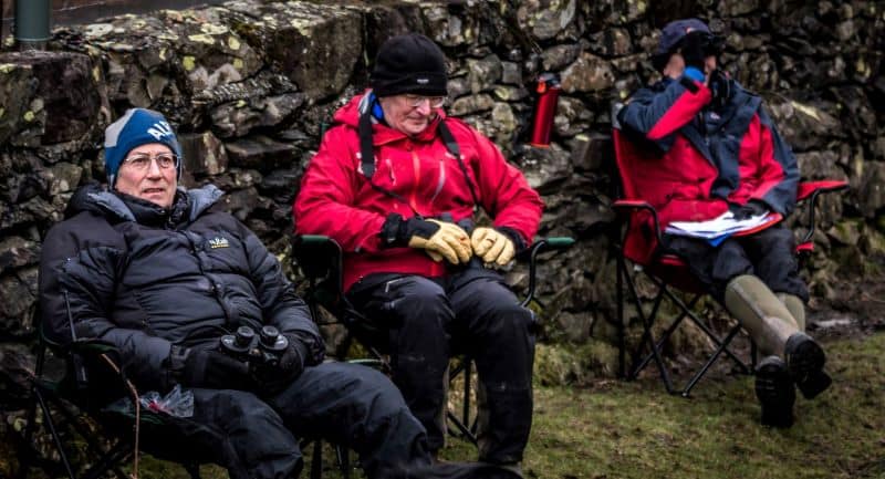 3 men sit in camp chairs with binoculars to observe the teams