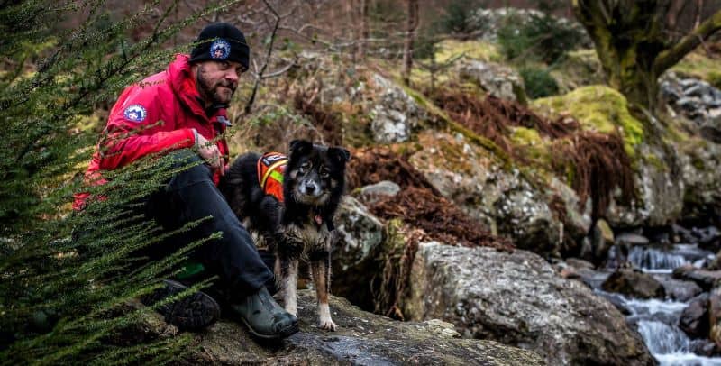 A dog handler kneels near a stream with a mixed breed search dog