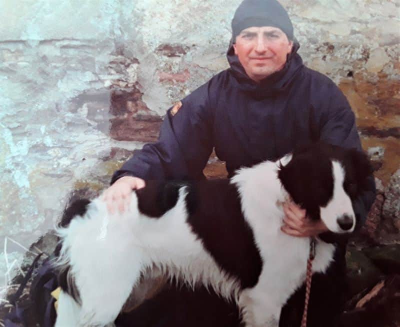 A man in a winter coat and hat poses beside a large black and white Border Collie.