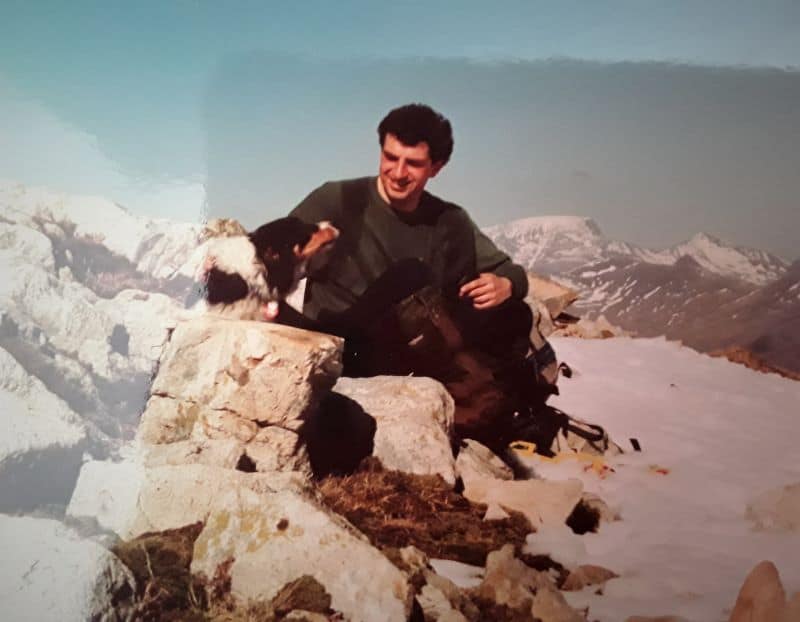 A young Stephen with curly dark hair sits on a snowy hillside with his tricolor Border Collie