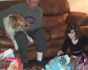 Charlie sits on a couch with Pixie in his lap while Isobel sits on the floor opening presents