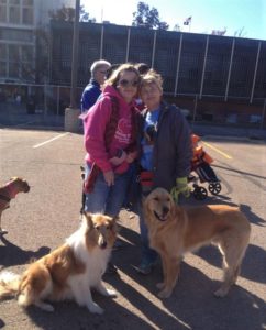 Two smiling women stand outside with a sable and white Collie and a Golden Retriever