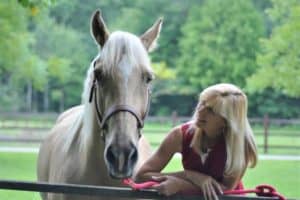 A blonde woman stands beside and smiles adoringly at a palomino horse