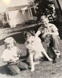 Black and white photo of a Rough Collie sitting outside between 2 kneeling children, one young girl and an older boy