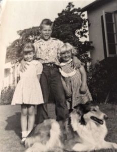 Vintage black and white photo of a sable and white Rough Collie lying at the feet of 3 children, one boy standing with his arms around the shoulders of 2 younger girls