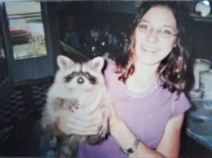 A brown-haired, smiling teenage girl holds a raccoon kit who is reaching for the camera