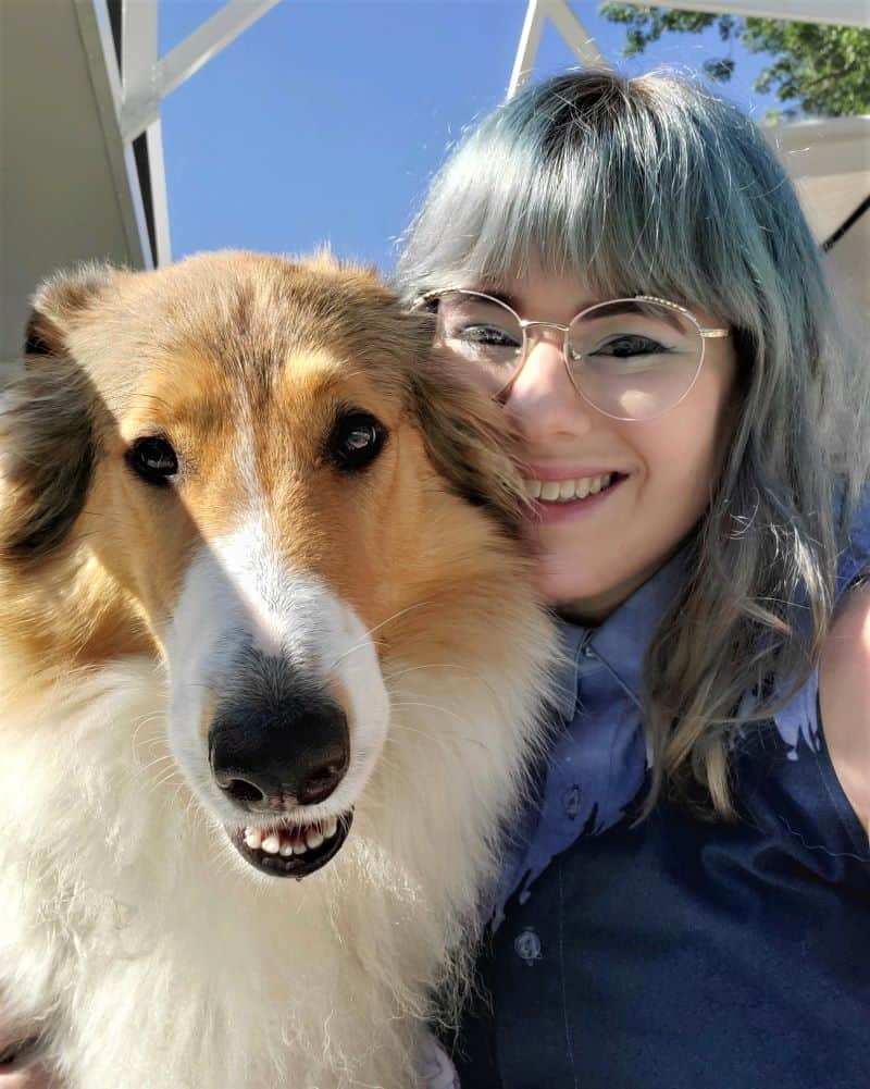 A sable-headed white Rough Collie (mostly white with tan markings on the head) smiles in a selfie with a smiling girl with blue/gray hair and glasses