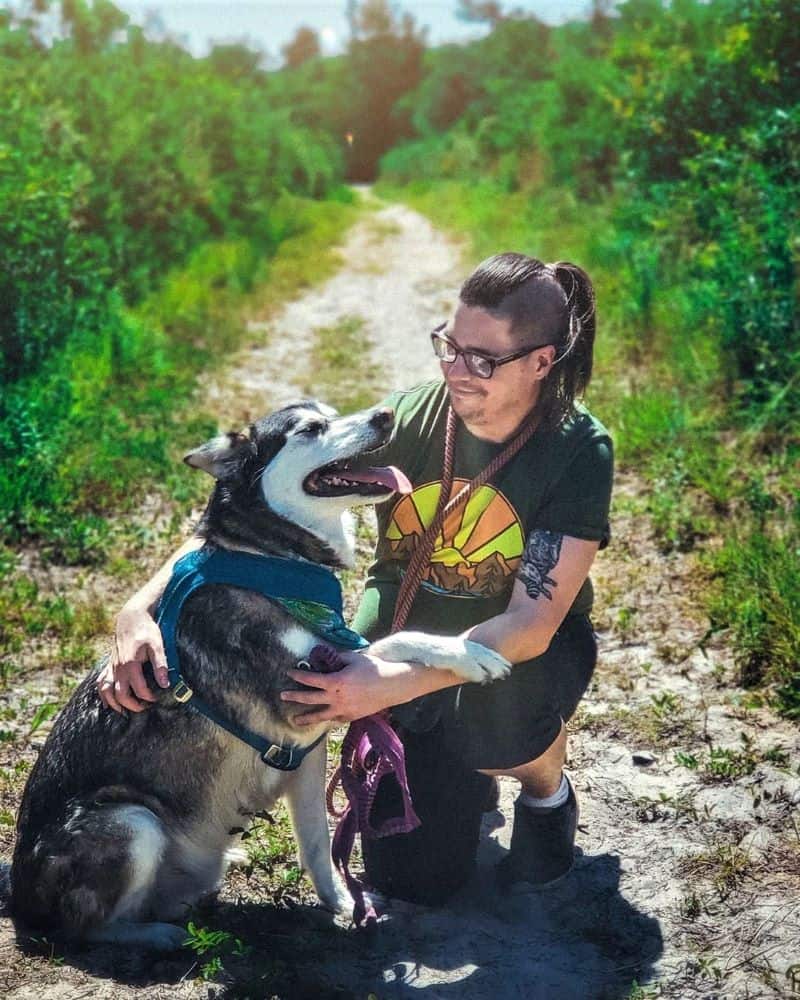 Dylan, a man with dark hair shaved on the sides and in a topknot ponytail, kneels on a wooded trail next to gray and white NAID Lakota