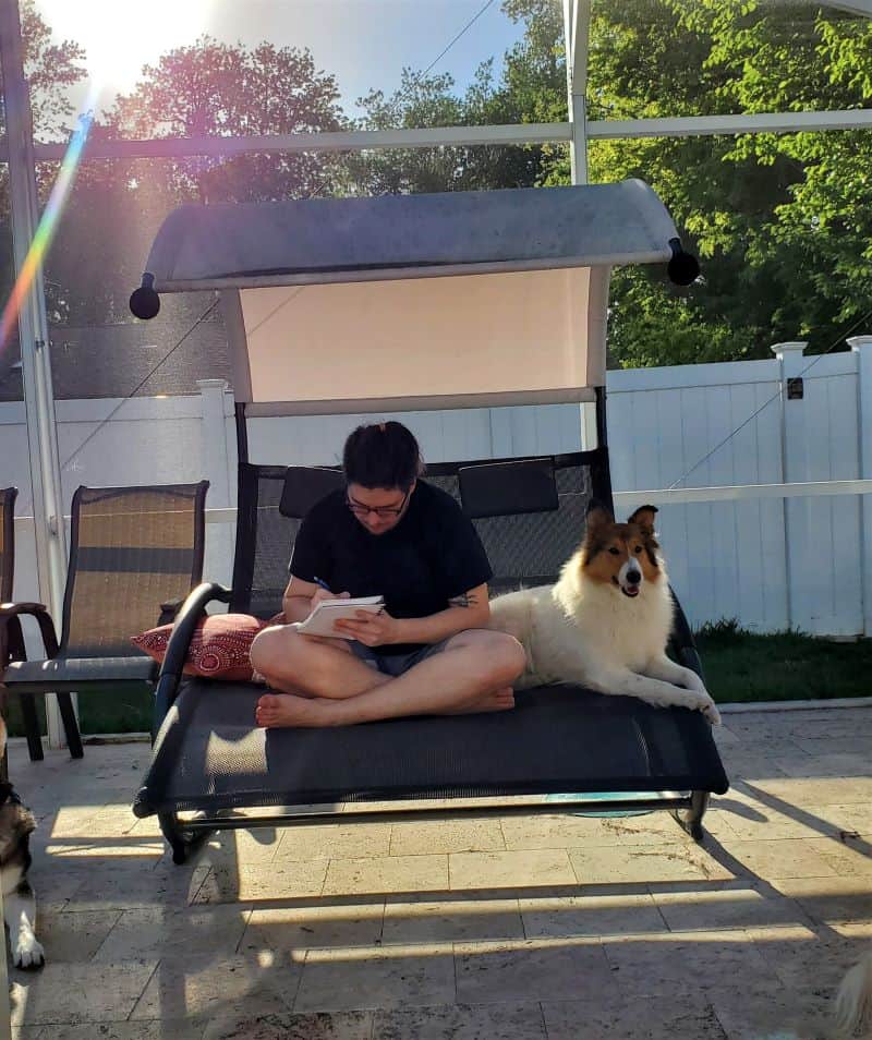 Vasya lies outside on a porch swing next to Dylan, who is hunched over diligently scribbling in a notebook