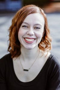 Headshot of Brianna smiling at the camera, her shoulder-length curly hair looking very red in the sunshine