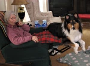 Pepsi has his hind feet on the floor and his front half draped across the footrest of a recliner while Grandma sips out a of a mug and smiles