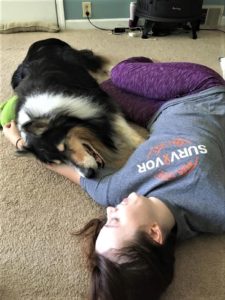 Pepsi lies on a carpeted floor beside Brianna, who is holding a toy in her hand, as if they flopped down to rest mid-play session