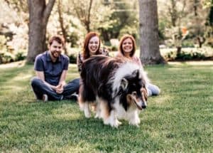 Pepsi walks away shaking his head while his human family, Pat, Brianna, and Brianna's mom sit on the grass behind him laughing at his antics