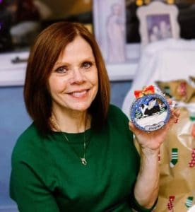 Brianna's mom, smiling, teary-eyed, and wearing a green shirt, holds up a personalized Christmas ornament of a tricolor Rough Collie crafted with wings that has Pepsi's name on the bottom