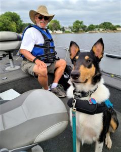 A tricolor Smooth Collie wearing a life vest rides contentedly in a boat next to an older man in a hat