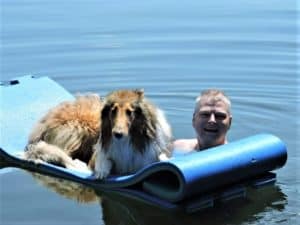 A sable and white Rough Collie lies on a large flotation device while his human pushes him through the lake