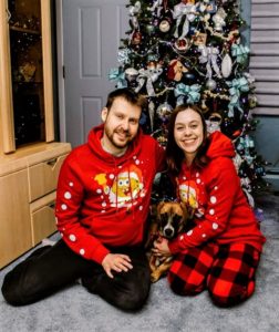A young couple sits before a Christmas tree wearing Sponge Bob Christmas pajamas and holding a Boxer mix puppy between them