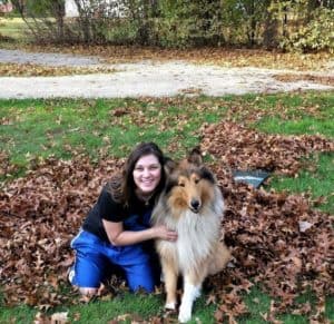 A sable and white Rough Collie and a girl wearing blue basketball shorts sit beside one another in a pile of leaves next to an abandoned garden rake