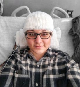 A girl with green eyes and glasses is in a hospital bed with her head wrapped in gauze and wires hanging down on the side