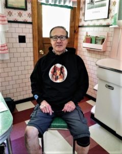 a man in glasses sits in a kitchen chair wearing a black hoodie with a sable and white Rough Collie's face on it