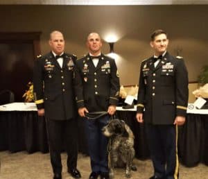 three military men in dress uniform pose for a picture, while the middle man holds the leash of a wirehaired german shorthair pointer