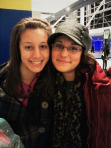 two girls, one wearing a headband with light brown hair and one wearing a hat with dark brown hair, smile at the camera together in an airport